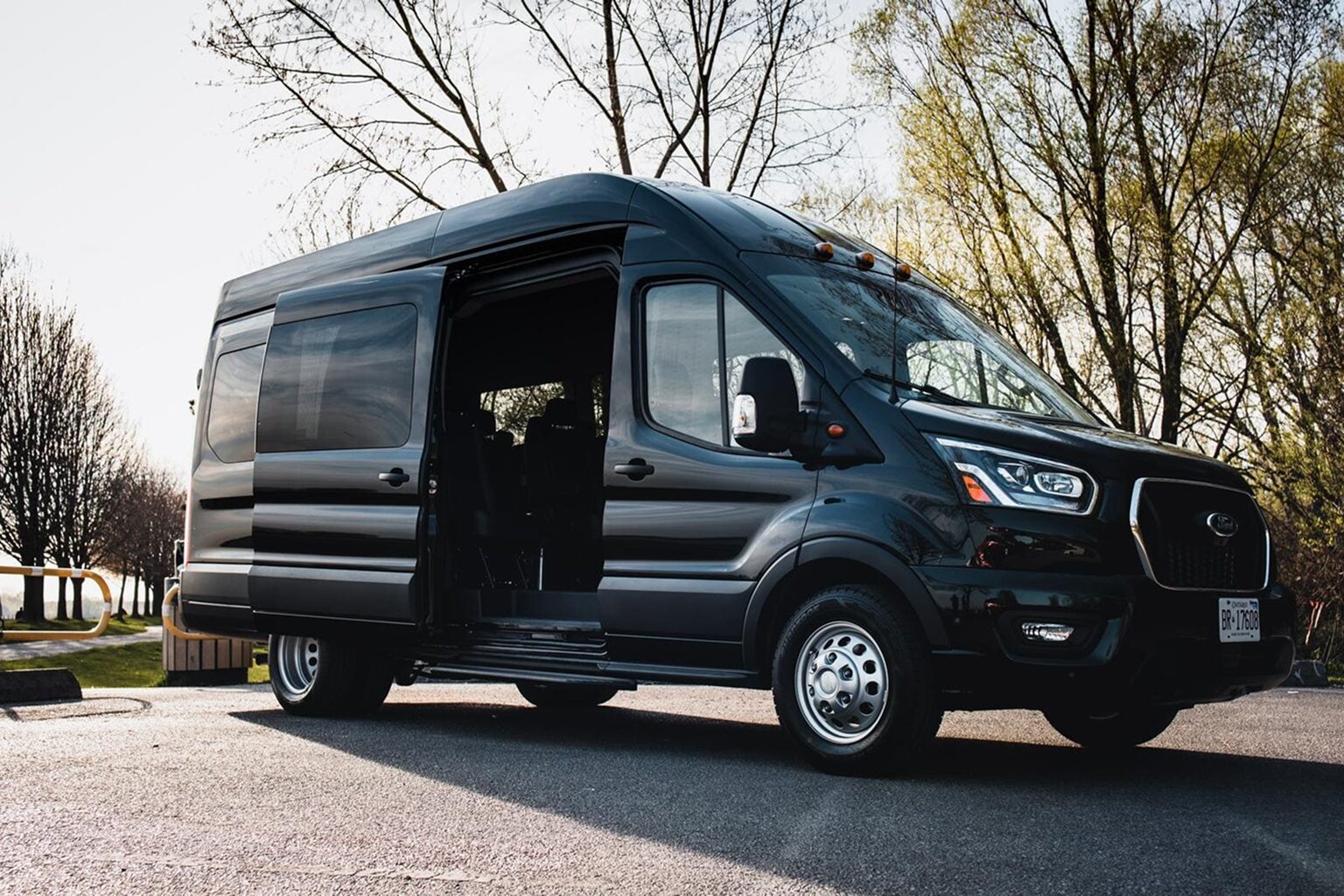 Black Ford Transit passenger van with open side door and trees in background.